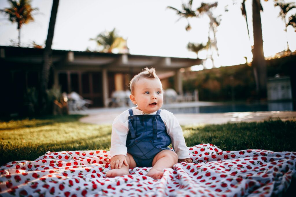 Cute baby sitting on a patterned blanket in a lush backyard during a sunny day.