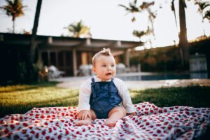 Cute baby sitting on a patterned blanket in a lush backyard during a sunny day.